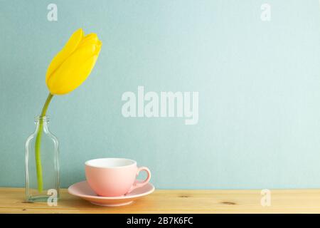 Coffee cup with yellow tulip flower in glass bottle on wooden table with mint green background. Floral arrangement, copy space Stock Photo