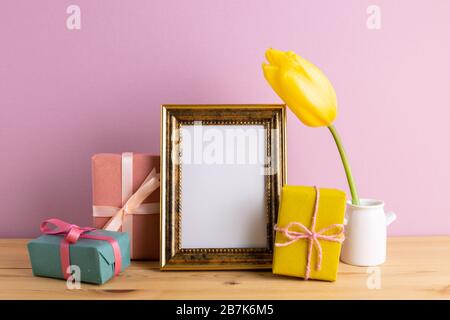 Photo frame with gift boxes and yellow tulip on wooden table with pink background. Anniversary concept, floral arrangement, copy space Stock Photo