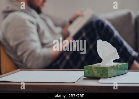 Box of tissues on the table, man on the background sitting on a sofa, reading a book. Sick at home concept. Stock Photo