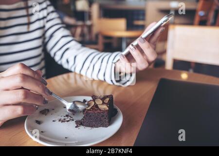 Closeup image of a woman using and looking at smart phone while eating brownie cake in cafe Stock Photo