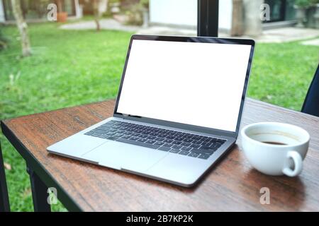 Mockup image of laptop with blank white desktop screen and coffee cup on wooden table with green nature background Stock Photo