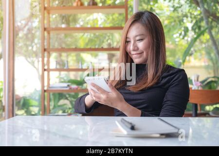 Closeup image of a beautiful business woman holding and writing on notebook in cafe with green nature background Stock Photo