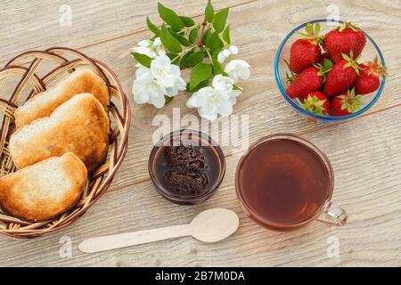 Glass cup of tea, homemade strawberry jam, fresh strawberries, spoon, toasts in wicker basket and white jasmine flowers on wooden background. Top view Stock Photo