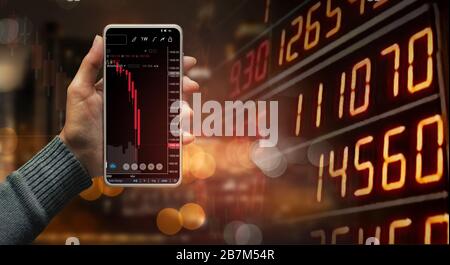 A hand holding a black smartphone showing stock market candlestick trend on blurred-out stock market data display panel, Business and finance concept. Stock Photo