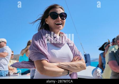 Boat trip on small ship on bay. Mature female with group of tourists enjoying sea voyage, sunny summer day, picturesque landscapes on horizon Stock Photo