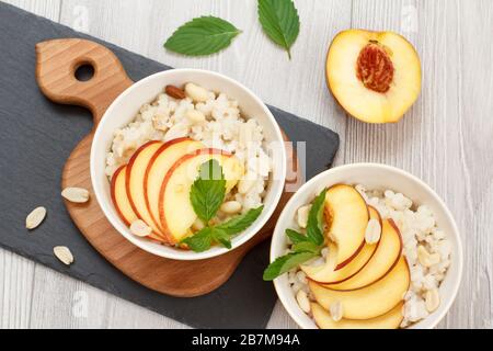 Sorghum porridge with pieces of peach, cashew nuts and almond in porcelain bowls, fresh peaches on wooden boards. Vegan gluten-free sorghum salad with Stock Photo