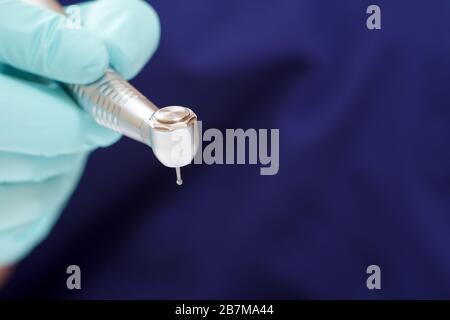 Dentist's hand in a green latex examination glove with new high-speed dental handpiece and blurred blue medical gown on the background. Medical tools Stock Photo