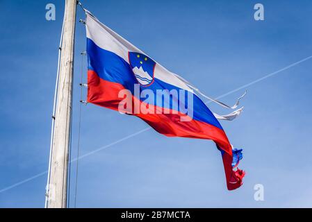 Torn and worn out Slovenian flag waving in the wind. Damaged, worn out, ripped fabric of Slovenia flag is waving in strong winds with blue skies and c Stock Photo