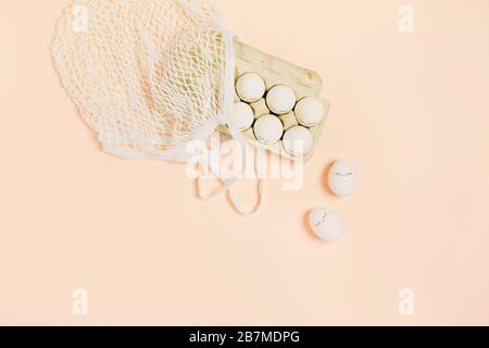 Happy Easter. White eggs with painted eyes in paper packaging and white mesh bag on peach color background. Flat lay, top view, copy space. Stock Photo