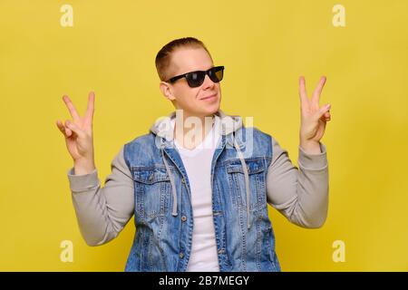 Stylish Caucasian man in a jeans and sunglasses on a yellow background. Hands abstractly shows the peace smiling. Close up. Stock Photo