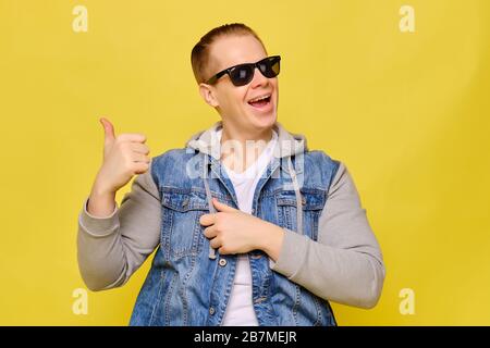 Stylish Caucasian man in a jeans and sunglasses on a yellow background. Copy space. Close up. Hands abstractly points a finger at an object smiling. Stock Photo