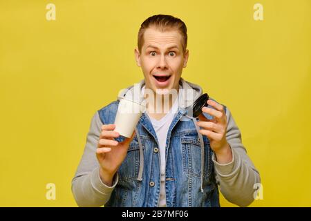 Stylish Caucasian man in jeans on a yellow background. Hands holding a disposable paper cup for coffee open and looking at the camera. Close up. Stock Photo