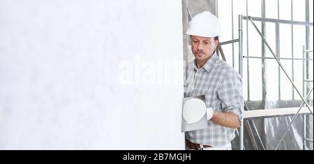 plasterer man at work with trowel plastering the wall of interior construction site wear helmet and protective gloves, scaffolding on background and c Stock Photo