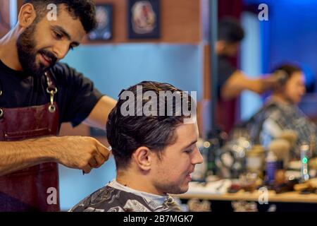 Master cuts hair and beard of men in the barbershop, hairdresser makes hairstyle for a young man Stock Photo