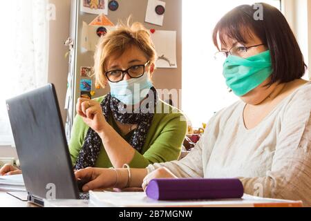 Two middle-age women teachers wearing surgical face mask discussing digital education at home during coronavirus restrictions Stock Photo