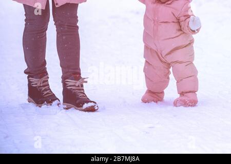 First steps. Mom holds the baby by the hand. Mom and little daughter walk in the snow in winter. The child takes his first steps outdoors in the snow Stock Photo