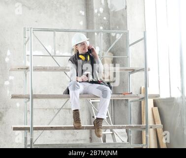 woman worker builder sitting on scaffolding to coffee break talking on mobile phone, wearing helmet, glasses and ear protection headphones, constructi Stock Photo