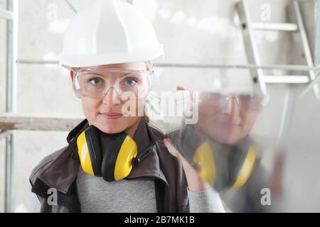 woman builder work with the straightedge spirit level, wearing helmet, safety glasses and hearing protection headphones, scaffolding interior construc Stock Photo