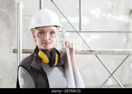 woman builder work with the straightedge spirit level, wearing helmet, safety glasses and hearing protection headphones, scaffolding interior construc Stock Photo