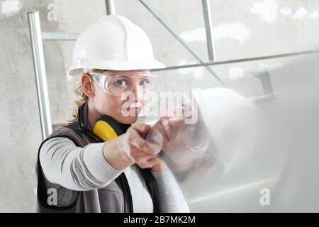woman builder work with the straightedge spirit level, wearing helmet, safety glasses and hearing protection headphones, scaffolding interior construc Stock Photo