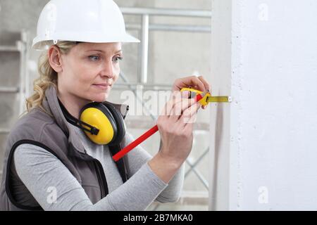 woman construction worker work with meter tape and pencil, measure wall in interior building site, wearing helmet and hearing protection headphones Stock Photo