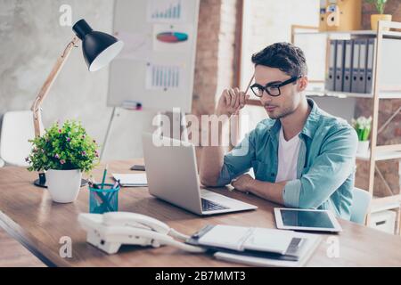 Young smart guy in jeans shirt and spectacles holding a pencil in his hand and trying to focus on the test Stock Photo