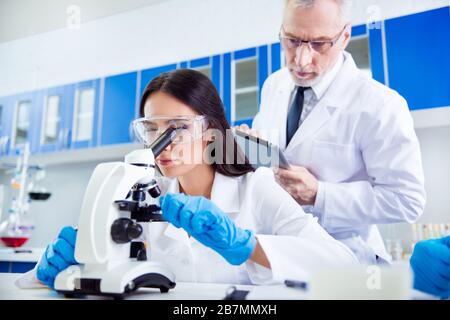 Laboratory, biotechnology, team work. Researcher is checking the esample on microscope and older professor is controlling her work and making notes Stock Photo