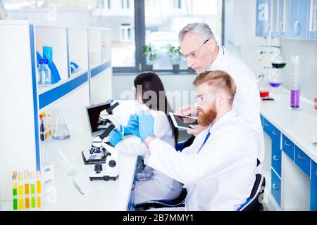 Researchers in labcoats are checking the samples on microscope in bright lab and older professor is controlling them and making notes on laptop. Great Stock Photo