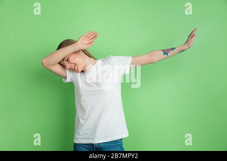 Winner's dab, dabbing. Caucasian young woman's portrait on green studio background. Beautiful female model in white shirt. Concept of human emotions, facial expression, sales, ad, youth. Stock Photo