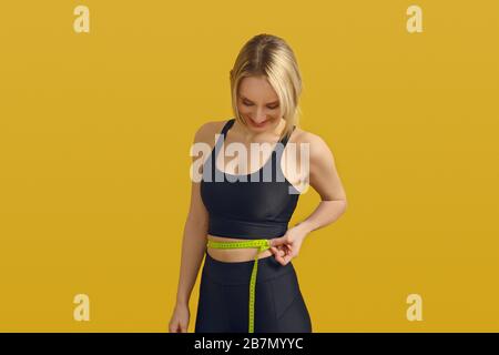 Slim sporty young woman measuring her waist with a tape measure as she looks down with a satisfied smile in a weight loss, healthy diet and exercise c Stock Photo