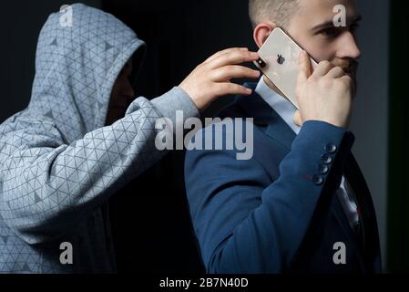 Zaporozhye. Ukraine. February 2020. Close-up portrait of a young bearded guy in a business suit holding an iPhone XS max. A robber in a dark hood, try Stock Photo