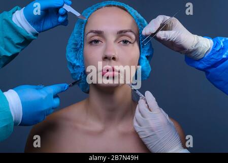 Close-up studio portrait of a young beautiful blonde girl, in a medical cap, next to four hands of plastic surgeons holding surgical instruments near Stock Photo