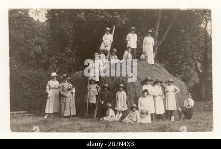 Early 1900's postcard of schoolgirls on country outing at harvest time, posing with a farmer and rakes, next to a haystack, circa 1923, U.K. Stock Photo