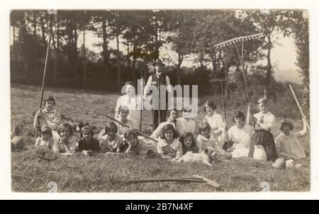 Early 1900's postcard of schoolgirls on country daytrip, posing with a farmer and rakes, circa 1923, U.K. Stock Photo