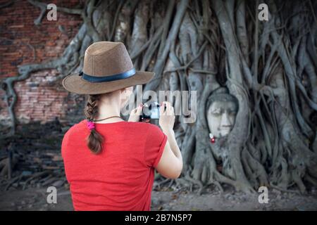 Woman tourist taking picture of Buddha head in Bodhi Tree Roots in Wat Mahathat complex in Ayutthaya, Thailand Stock Photo