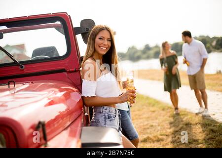 Happy young women drinks cider from the bottle by the convertible car with friends Stock Photo