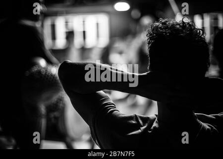 A close up selective focus atmospheric shot in black and white. A muscular man relaxes indoor, with hands behind head. Male silhouette from behind. Stock Photo
