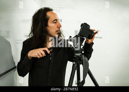 A closeup head profile shot of a caucasian man with long black hair using a camera mounted on a tripod during a filming video session in studio. Stock Photo