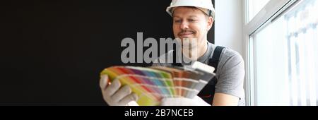 Young repairman in helmet sitting in flat and choosing color for interior Stock Photo