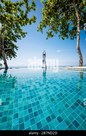 A young woman practicing yoga under palm trees next to an infinity pool Stock Photo