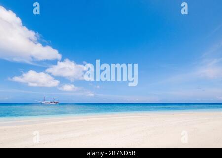 A tall masted ship moored on a tropical ocean in front of a pristine beach Stock Photo