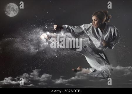 Young impudent emotional karate girl in a suit jumps up and makes a powerful blow on the background of clouds, sky and moon. Unrestrained energy conce Stock Photo