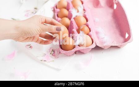 Close-up view of raw chicken eggs in egg box on white background.Hand in the frame. Decor and feathers. DEEP EXPOSURE, SELECTIVE FOCUS. Free space for text.  Stock Photo