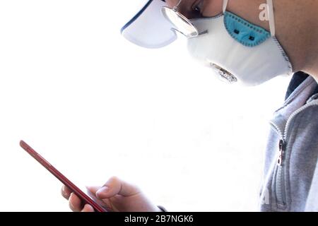 Side view of man face in a cap and glasses. A man holds a phone in his hand and looks at it. Medical respirator. White background with copy space. The Stock Photo