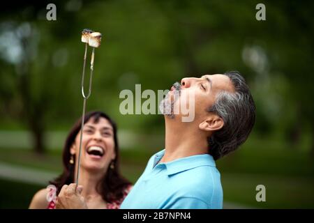 Middle-aged woman laughs in delight as her husband blows on the marshmallows he has burnt over the campfire. Stock Photo