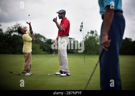 Mid adult man talking to his son as his son throws a golf ball in the air on a golf course. Stock Photo