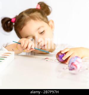 Beautiful small girl painted Easter eggs, time at home, paints and brushes on white table. Preparation for the Easter. Idea How Parents Can Keep Kids Busy in Coronavirus Quarantine as Schools Close. Stock Photo