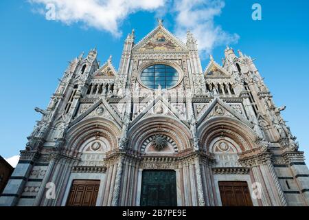 Looking up at decorated front exterior of Siena Cathedral. Intricate stonework, colorful religious imagery & blue sky reflected in rose window