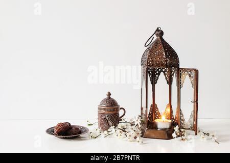 Ramadan Kareem greeting card, invitation. Bronze plate with dates fruit, white flowers, prunus tree blossoms, cup of tea and glowing Moroccan lantern Stock Photo