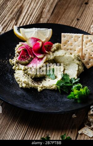 Healthy snack, gourmet dip. Green hummus with pickled watermelon radishes and lemon slices on top in black ceramic bowl on rustic wooden table Stock Photo